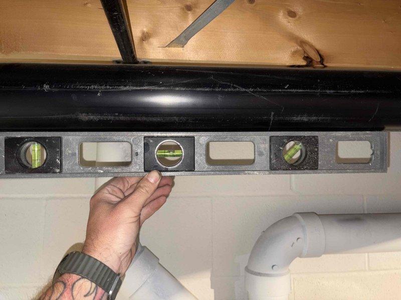 Responded to callback for water leakage from drainage pipe onto shelving unit. Upon inspection, verified the black CORE DWV (Drain-Waste-Vent) flexible pipe installation for proper pitch and alignment using a spirit level. Examined pipe connections and clamping points where sections join together against the foundation wall. Documented existing conditions showing visible wear, debris accumulation, and potential points of water seepage on the corrugated drainage line. Assessed the horizontal run of the DWV pipe secured to ceiling joists above the affected area to identify source of dripping water reported by customer.
